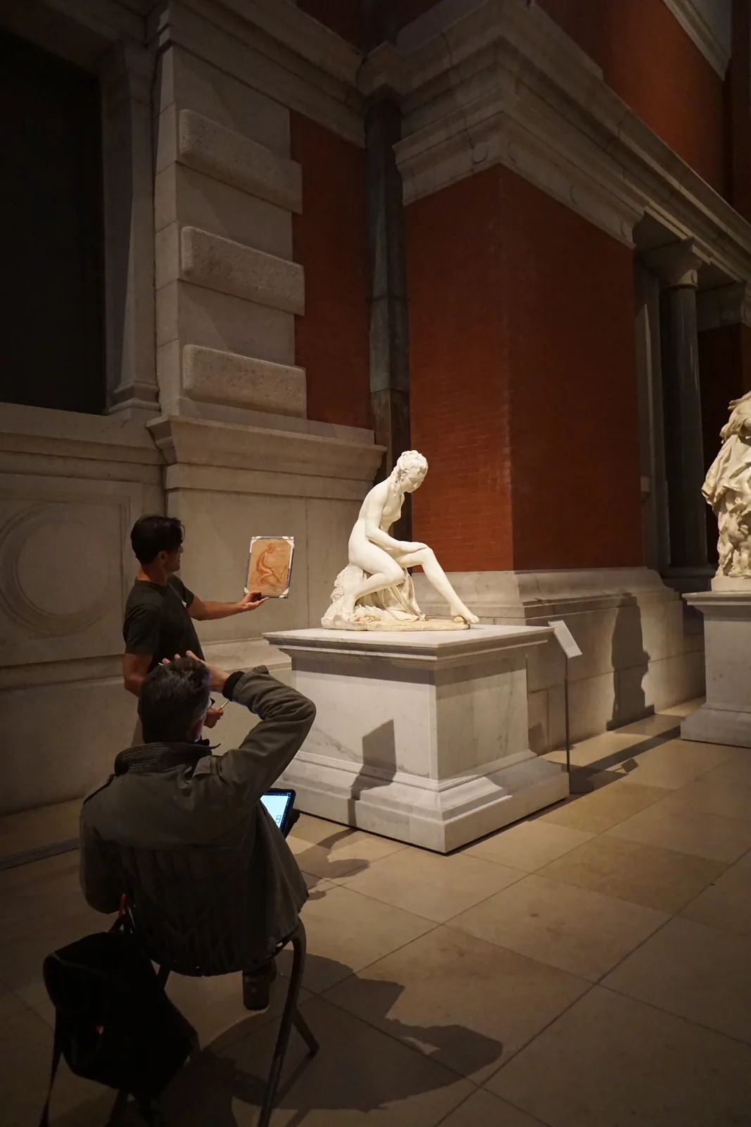 Two visitors drawing a marble sculpture in a museum gallery, one seated with a tablet